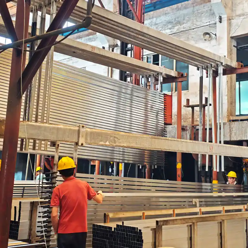 Factory workers securing long aluminum profiles onto vertical racks in preparation for the anodizing process.